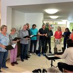 Ein Foto vom Auftritt im Seniorenzentrum. Der Chor Cantiamo steht im Halbkreis vor seiner Chorleiterin, die den Chor sitzend am Klavier begleitet.