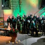 Das Foto zeigt den Chor Cantiamo bei seinem Konzert "Eine Reise um die Welt" in der evangelischen Kirche Grünwettersbach. Vor dem Chor sitzt eine Pianistin, begleitet wird sie von einer Violinistin. Auf dem Taufbecken vor dem Chor liegt ein Adventskranz, auf dem zwei Kerzen brennen.