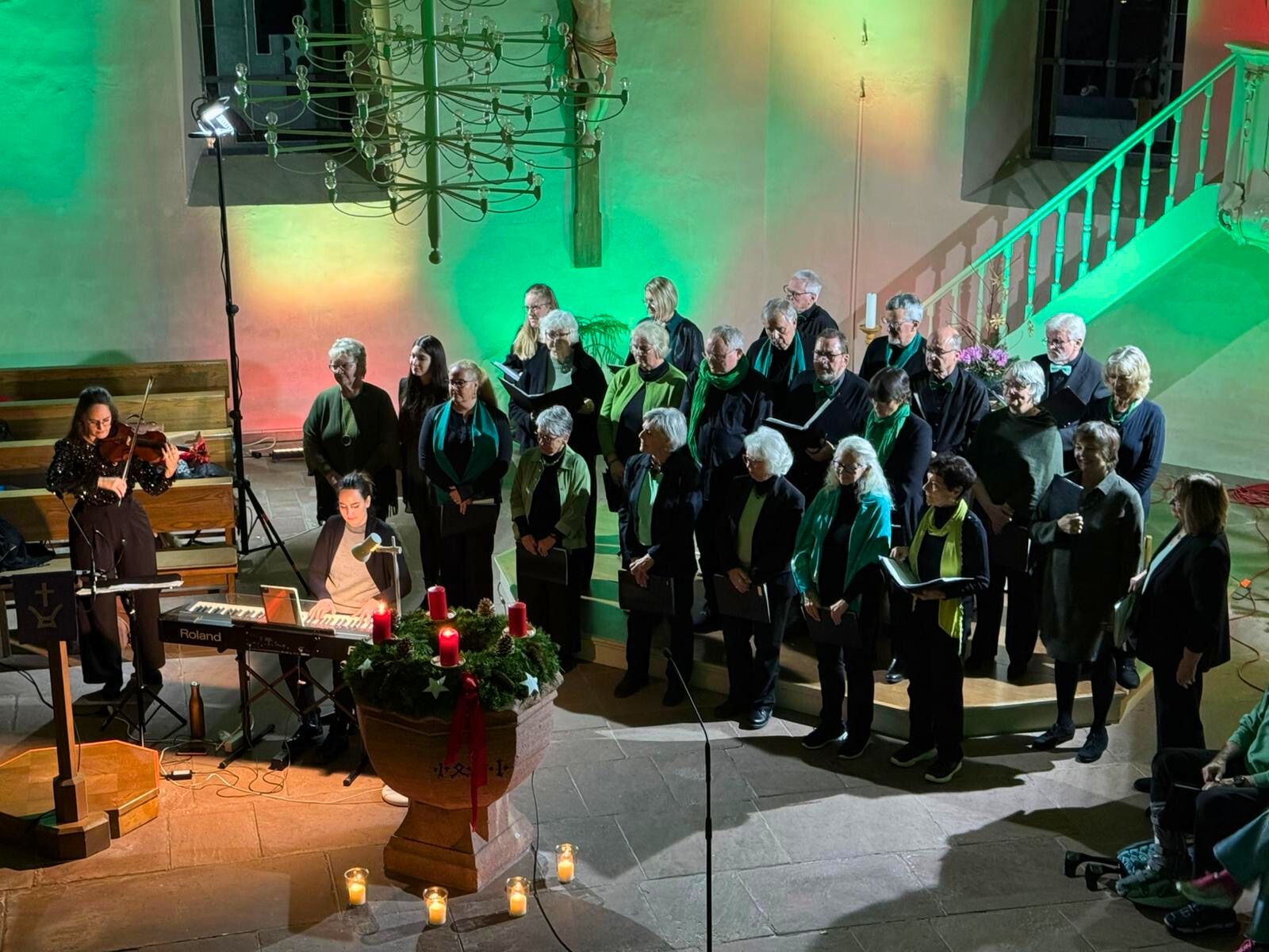 Das Foto zeigt den Chor Cantiamo bei seinem Konzert "Eine Reise um die Welt" in der evangelischen Kirche Grünwettersbach. Vor dem Chor sitzt eine Pianistin, begleitet wird sie von einer Violinistin. Auf dem Taufbecken vor dem Chor liegt ein Adventskranz, auf dem zwei Kerzen brennen.