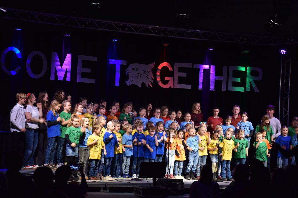 Ein Foto des Come Together-Konzerts vom 15. November 2025. Die bunt gekleideten Kinder des Kinderchores "SingKids" stehen auf einer Bühne. Vor ihnen sind Umrisse des Publikums und des Dirigenten zu sehen. Hinter ihnen hängt groß und bunt angestrahlt der Schriftzug "Come Together".