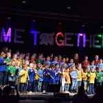 Ein Foto des Come Together-Konzerts vom 15. November 2025. Die bunt gekleideten Kinder des Kinderchores "SingKids" stehen auf einer Bühne. Vor ihnen sind Umrisse des Publikums und des Dirigenten zu sehen. Hinter ihnen hängt groß und bunt angestrahlt der Schriftzug "Come Together".