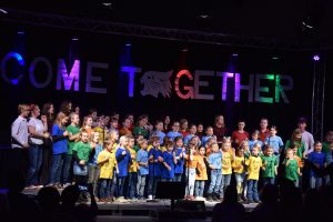 Ein Foto des Come Together-Konzerts vom 15. November 2025. Die bunt gekleideten Kinder des Kinderchores "SingKids" stehen auf einer Bühne. Vor ihnen sind Umrisse des Publikums und des Dirigenten zu sehen. Hinter ihnen hängt groß und bunt angestrahlt der Schriftzug "Come Together".