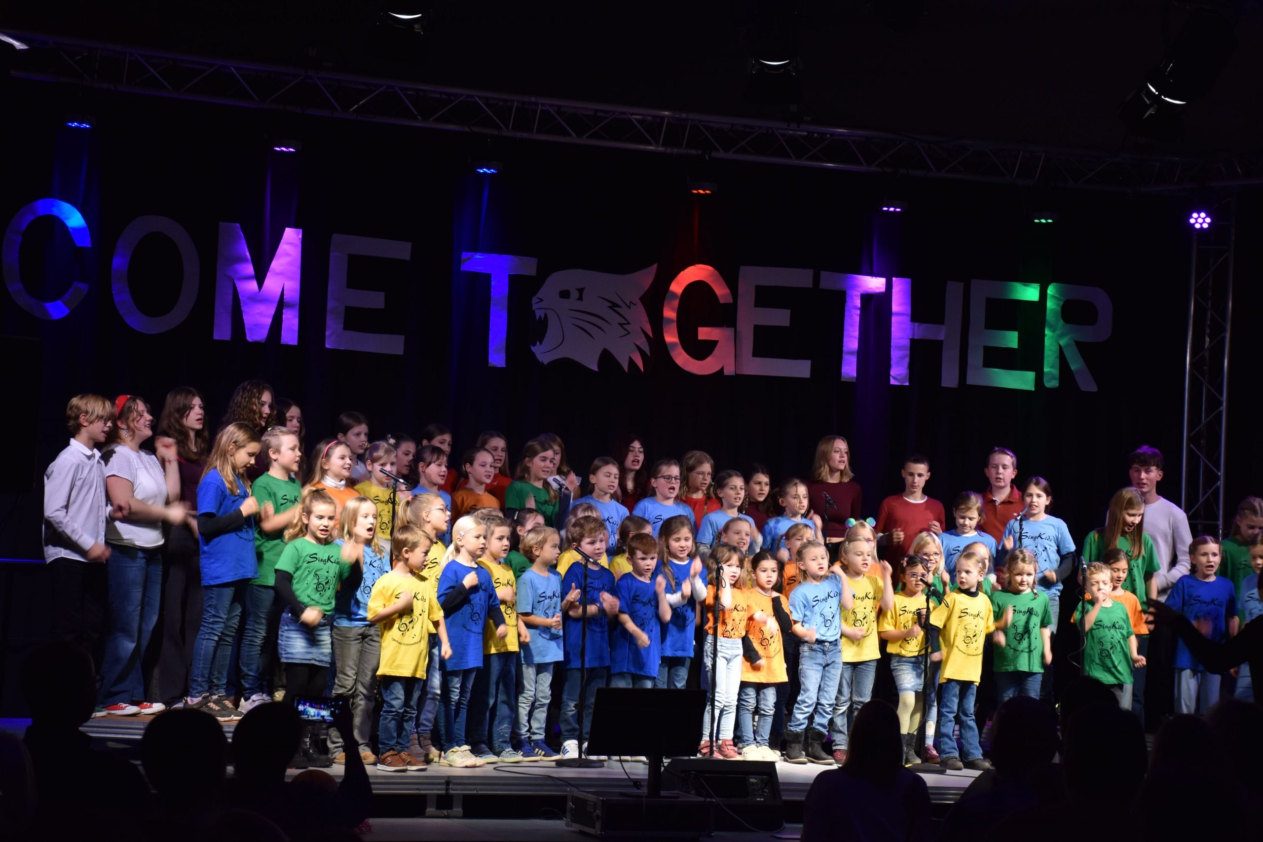 Ein Foto des Come Together-Konzerts vom 15. November 2025. Die bunt gekleideten Kinder des Kinderchores "SingKids" stehen auf einer Bühne. Vor ihnen sind Umrisse des Publikums und des Dirigenten zu sehen. Hinter ihnen hängt groß und bunt angestrahlt der Schriftzug "Come Together".
