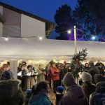 Das Foto zeigt den PopChor des Sängerhains Grünwettersbach bei seinem Auftritt auf dem Adventsmarkt auf dem Pfeilerhof. Der Chor steht von weihnachtlichen Lichterketten in warmes Licht getaucht in einem Festzelt. Im Vordergrund sind Besucher:innen des Adventsmarkts sowie Chorleiter Tobias Thimig am E-Piano zu sehen.