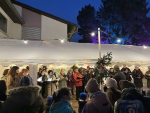 Das Foto zeigt den PopChor des Sängerhains Grünwettersbach bei seinem Auftritt auf dem Adventsmarkt auf dem Pfeilerhof. Der Chor steht von weihnachtlichen Lichterketten in warmes Licht getaucht in einem Festzelt. Im Vordergrund sind Besucher:innen des Adventsmarkts sowie Chorleiter Tobias Thimig am E-Piano zu sehen.